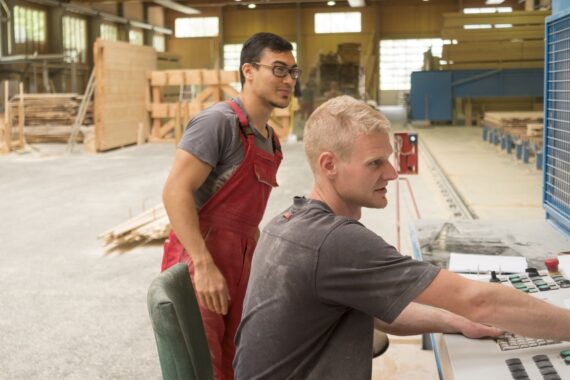 Das Bild zeigt zwei Mitarbeiter bei der Arbeit im Thoma Holz100 Werk in Stadl. Das Bild ist Teil einer Stellenanzeige für Maschinenführer*innen. - Interesse an einem Job bei uns ? - Thoma Holz100